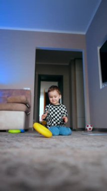 Kareli tişörtlü çocuk odada yerde oturuyor. Mutlu çocuk oyuncaklarla oynar ve kameraya doğru koşar. Düşük açı. Dikey video.