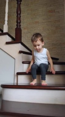 Küçük beyaz çocuk, merdivenlerde içeride oturuyordu. Bebek ayağa kalkar ve uzaklaşır. Dikey video.