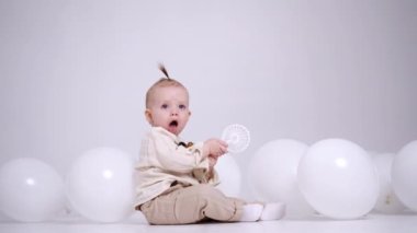 Küçük sevimli beyaz bebek stüdyoda, beyaz balonların arasında yerde oturuyor. Sevimli çocuk oyuncaklarla oynar ve onu ağzına alır..