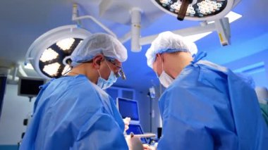 Mavi üniformalı ve cüppeli iki erkek doktor aletleri tutuyor. Cerrahların ameliyata başlaması. Arka planda yuvarlak lambalar.