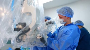 Mavi üniformalı birkaç doktor ameliyathanede çalışıyor. Tıbbi asistanlar ameliyat öncesi ekipmanları plastik kaplamayla düzenlerler..