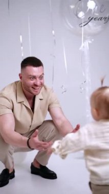 Gülümseyen baba ellerini öne uzatarak çömelmiş oturuyor. Küçük bebek yaklaşıyor ve baba çocuğu kucağına alıyor ve sevgiyle öpüyor. Dikey video.
