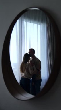Evdeki pencerenin önünde duran sevgi dolu bir ailenin yansıması. Birbirlerine yakın durup bebek tutuyorlar. Adam karısını alnından öpüyor. Dikey video.