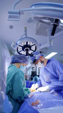 İki doktor ameliyathanede ameliyat yapıyor. Cerrahların üstündeki tavanda yuvarlak lambalar. Dikey video.