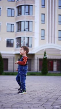 Kot pantolon giyen beyaz bir çocuk dışarıda dikiliyor. Bebek ona doğru uçan sabun köpüklerine bakıyor. Dikey video.