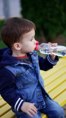 Siyah saçlı erkek bebek caddedeki bankta oturuyor. Huzurlu çocuk şişeden içiyor. Üst Manzara. Dikey video.
