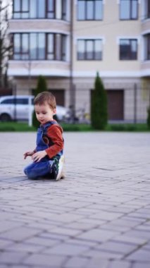 Kot pantolon giyen beyaz bir çocuk açık havada, yerde oturuyordu. Çocuk dışarıda. Dikey video.