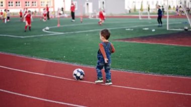 Küçük çocuk stadyumun etrafında yuvarlanan topun peşinden koşuyor. Bebek çocuk, arka planda çocuk futbol takımına bakmak için duruyor..