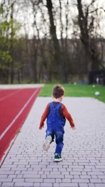 Asfalt zeminde koşan gömlek ve kot pantolon giyen bir bebeğin arka manzarası. Toddler topu yakalamak için koşuyor. Dikey video.