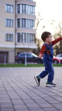 Kafkasyalı çocuk neşeyle dışarı koşuyor. Sabun köpüklerini yakalayan güzel bebek. Dikey video.