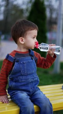 Koyu renk saçlı, kot pantolon giyen beyaz çocuk şişeden su içiyor. Sevimli bebek dışarıda bankta oturuyor. Dikey video.