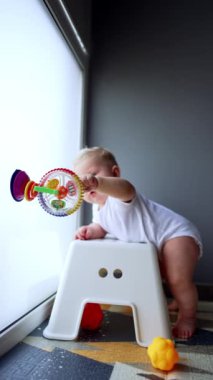 Beyaz tulum giyen sevimli erkek bebek sandalyeye yaslanıyor. Pencereye yapışmış bir oyuncakla sevimli bir bebek oynuyor. Dikey video.