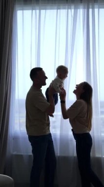 Pencerenin önünde duran küçük, sevimli oğlunu tutan adam. Sevgi dolu anne perdelerin arkasında saklanan bebekle oynar. Dikey video.