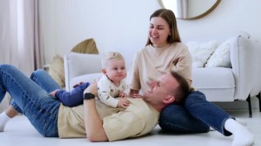Küçük sevimli sarışın bebek babasının karnında yatıyor. Yanında oturan anne oğluna sevgiyle bakıyor. Aile bir arada.