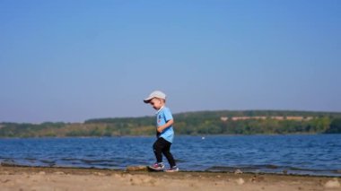 Erkek bebek güneşli bir günde nehir kıyısında yürür. Beyaz çocuk plajda taş arıyor..