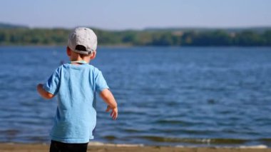 Nehre doğru koşan küçük bir çocuğun arka görüntüsü. Çocuk bir taş taşıyor ve suya atıyor..