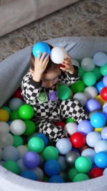 Kuru havuzda oturup plastik toplarla oynayan tatlı bir çocuk. Evde oynayan neşeli çocuğun en iyi görüntüsü. Dikey video.