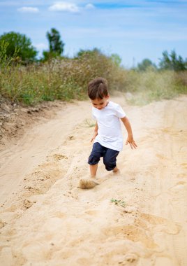 Kumlu yolda koşan bir çocuk dışarıda eğlenceli bir gün geçiriyor. Beyaz gömlekli genç bir çocuk kumlu bir patikada neşeli bir şekilde oynar, günün sıcaklığını hisseder..