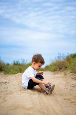 Genç bir çocuk bir yaz öğleden sonrası boyunca gün ışığında kumlu yolda oturuyor. Güneşli bir günde beyaz gömlekli bir çocuk kumlarda oynar.