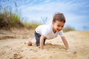 Küçük çocuk güneşli bir günde kumlu alanda neşeyle oynuyor. Genç bir çocuk mutlu bir şekilde kumda sürünüyor, neşeli anların tadını çıkarıyor.