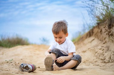 Güneşli bir günde kumsalda oynayan genç bir çocuk. Neşeli bir çocuk sıcak kumda oturur, güneşli bir plaj gününün tadını çıkarırken ayakkabısını keşfeder..