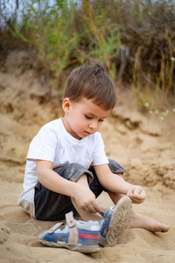 Genç bir çocuk kumsalda oturuyor, güneşli açık havada ayakkabıyla oynuyor. Genç bir çocuk, açık havada yumuşak kumda otururken ayakkabısına odaklanmış bir şekilde öne doğru eğiliyor.