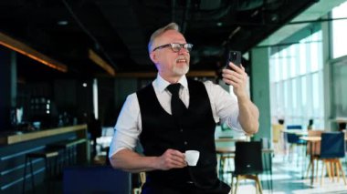 Sakallı adam kahve molası veriyor ve telefonunu kullanıyor. Pozitif adam selfie çekiyor ya da video çekiyor..