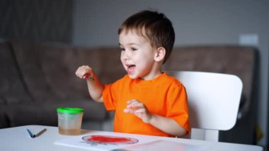 Bebek fırçayı fincanda yıkıyor ve neşeyle gülümsüyor. Annesinin eli çocuğunun resmine işaret ediyor. Yürümeye başlayan çocuk için sanat.