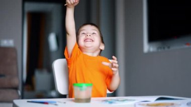 Turuncu tişörtlü güzel çocuk sanat aktivitesi yapıyor. Çocuk fırçayı yıkıyor ve sevinçle kaldırıyor. Bulanık arkaplan.