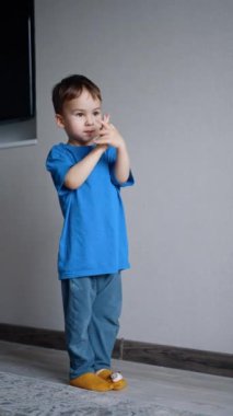 İki yaşında beyaz, şirin bir çocuk duvarın içinde dikiliyordu. Mavi tişört giyen bebek ellerini çırpıyor. Dikey video.