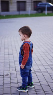 Koyu saçlı, beyaz erkek bebek dışarıda gömlek ve kot pantolon giyerek dikiliyor. Çocuk üç parmağını göstererek kameraya döndü ve kaçtı. Dikey video.