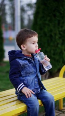 Bankta oturan sevimli çocuk şişeyi tutuyor. Çocuk şişeyi ağzında tutuyor ama içmiyor. Dikey video.