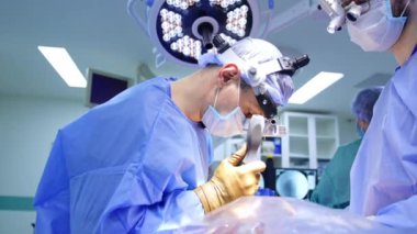 Beyaz erkek beyin cerrahı, vida sürücüsüne benzer bir alet kullanıyor. Ameliyat masası takan iki doktor ameliyatta çalışıyor..