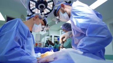 Cerrahi hoparlörlerde çalışan beyin cerrahı bir hastanın her iki tarafında da bulunur. Uzun doktor tornavidayı, diğerinin cerrahi alet kullandığı gibi kullanıyor. Hemşire arka planda.