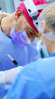 Operasyonda kep ve far takan profesyonel bir erkek cerrah. Doktor elektrikli cihaz kullanıyor. Kapatın. Dikey video.