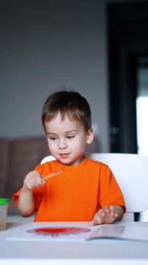 Turuncu tişört giyen konsantre olmuş üç yaşında bir çocuk gazetenin yanındaki fırçayı hareket ettiriyor. Tatlı çocuk fırçayı bardakta yıkıyor. Dikey video.
