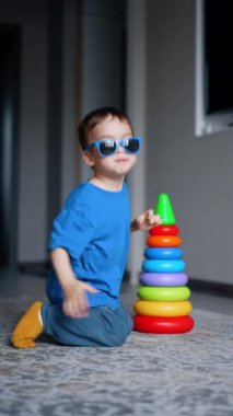 Güneş gözlüğü takan sevimli beyaz çocuk yerde piramitlerle oynuyor. Çocuk oyuncağı kaldırıyor ve kameraya yaklaşıyor. Düşük açı. Dikey video.