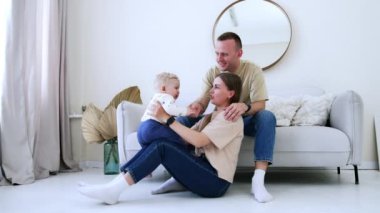 Sevimli küçük bebek annesinin kucağına oturuyor. Anne çocuğunu onu yukarı aşağı kaldırırken eğlendiriyor. Baba kanepeye oturup oğlunu neşelendiriyor..