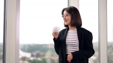 Pencerenin önünde duran beyaz kadın. Kadın pencereye bakarak kahve içiyor. Düşük açı görünümü.