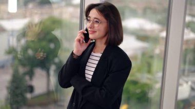 Gözlüklü ve siyah ceketli çekici beyaz kadın pencerede duruyor. Kadın telefonda gülümsüyor..