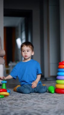 Mavi tişört ve pantolon giyen küçük çocuk oyuncaklarının arasında yerde oturuyor. Toddler ayağa kalktı ve kameraya yaklaştı. Dikey video.