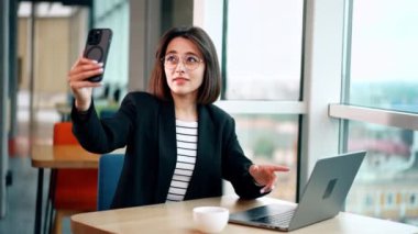 Siyah saçlı, gözlüklü beyaz kadın selfie çekiyor. Kadın dizüstü bilgisayarını çevirir ve dikkatlice bakar..