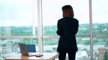 Panoramik pencerede duran koyu renk takım elbiseli bir kadının dikiz görüntüsü. Ofisteki işine ara veren bir bayan..