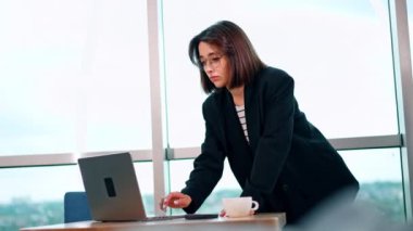 Masanın başında dizüstü bilgisayara yaslanmış beyaz bir kadın. Bayan gülümseyerek bilgisayarını kullanıyor. Düşük açı görünümü.