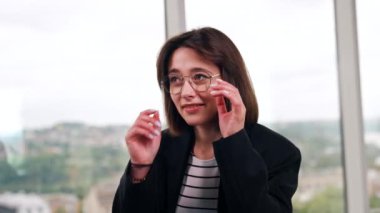 Siyah ceketli ve gözlüklü güzel beyaz esmer kadın. Bayan gözlüklerini çıkardı ve gülümseyen uydu kamerasına baktı. Portreyi kapat. Bulanık arkaplan.