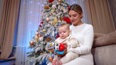 Beyaz bir kadın kucağında bebekle yerde oturuyor. Çocuk elindeki oyuncağa bakıyor. Noel ağacı arka planda. Düşük açı görünümü.