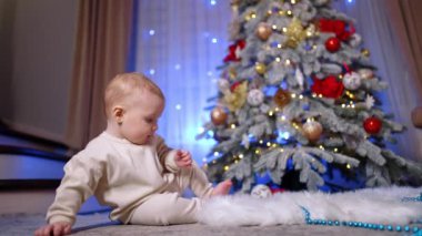Beyaz takım elbiseli güzel bir bebek Noel ağacının yanında yerde oturuyor. Sevimli sevimli çocuk kamerayı fark eder ve sürünerek yaklaşır..