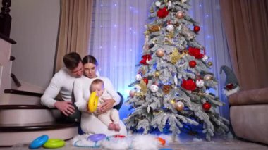 Mutlu ebeveynler ve bebekleri Noel ağacının yanındaki merdivenlerde oturuyorlar. Tatlı çocuk piramitlerle oynuyor..