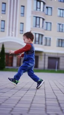 Enerjik mutlu beyaz çocuk sabun köpüğü yakalamak için koşuyor. Üç yaşında neşeli bir bebek açık havada vakit geçiriyor. Dikey video.
