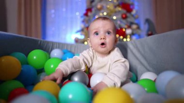 Hayretler içinde beyaz bir bebek kuru havuzda oturuyor. Sevimli çocuk etrafındaki plastik topa dokunuyor. Arkaplanda bulanık bir Noel ağacı.